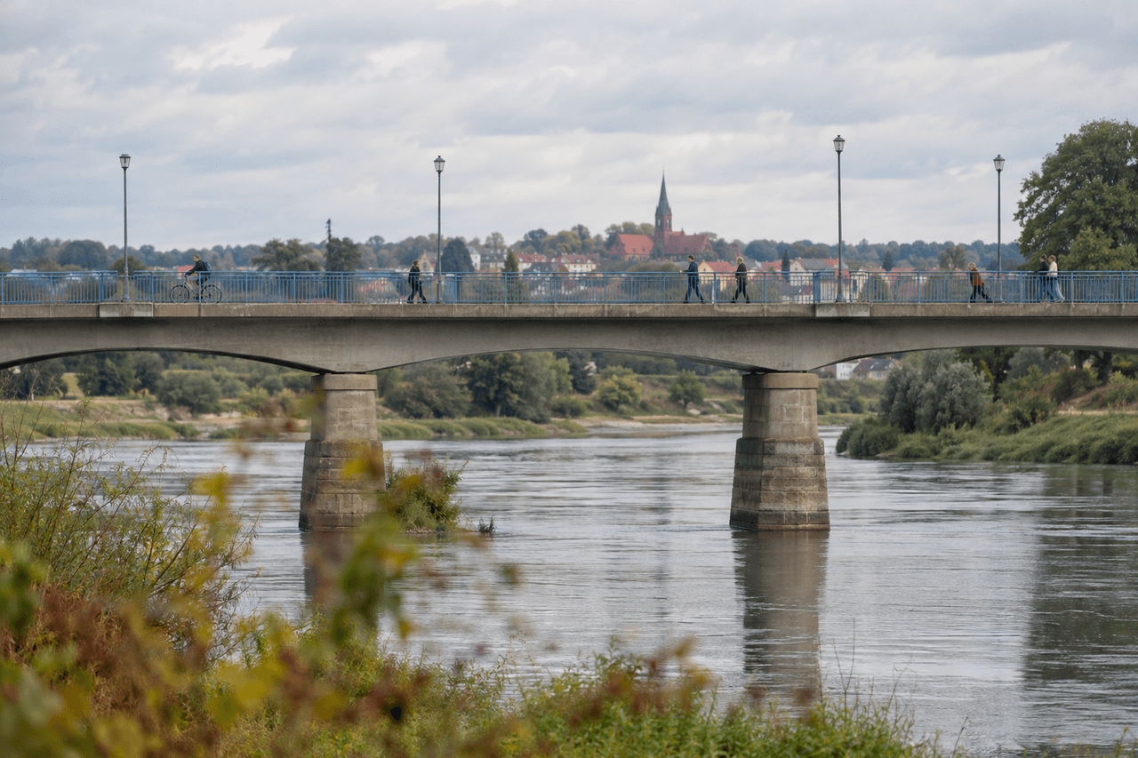 Fußgänger und ein Radfahrer überqueren an einem bewölkten Tag eine Brücke über einen breiten Fluss, dahinter eine Kleinstadt mit Kirchturm