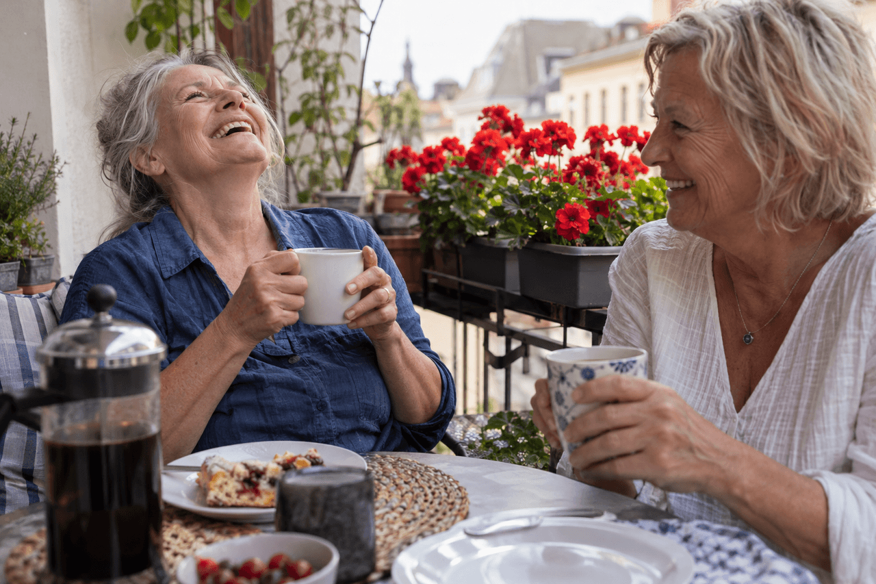 Zwei Frauen um die sechzig lachen beim Kaffeetrinken auf einem Balkon mit roten Geranien