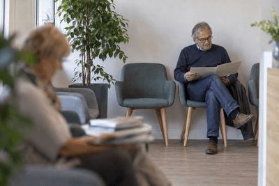 Mann sitzt im hellen Wartebereich einer Praxis und liest in einer Mappe, im Vordergrund unscharf eine wartende Frau
