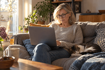 Frau mittleren Alters sitzt mit dem Laptop auf dem Sofa, neben ihr schläft eine Katze
